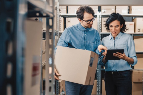 Man and woman standing in room with cardboard boxes on racking, looking at something on a tablet