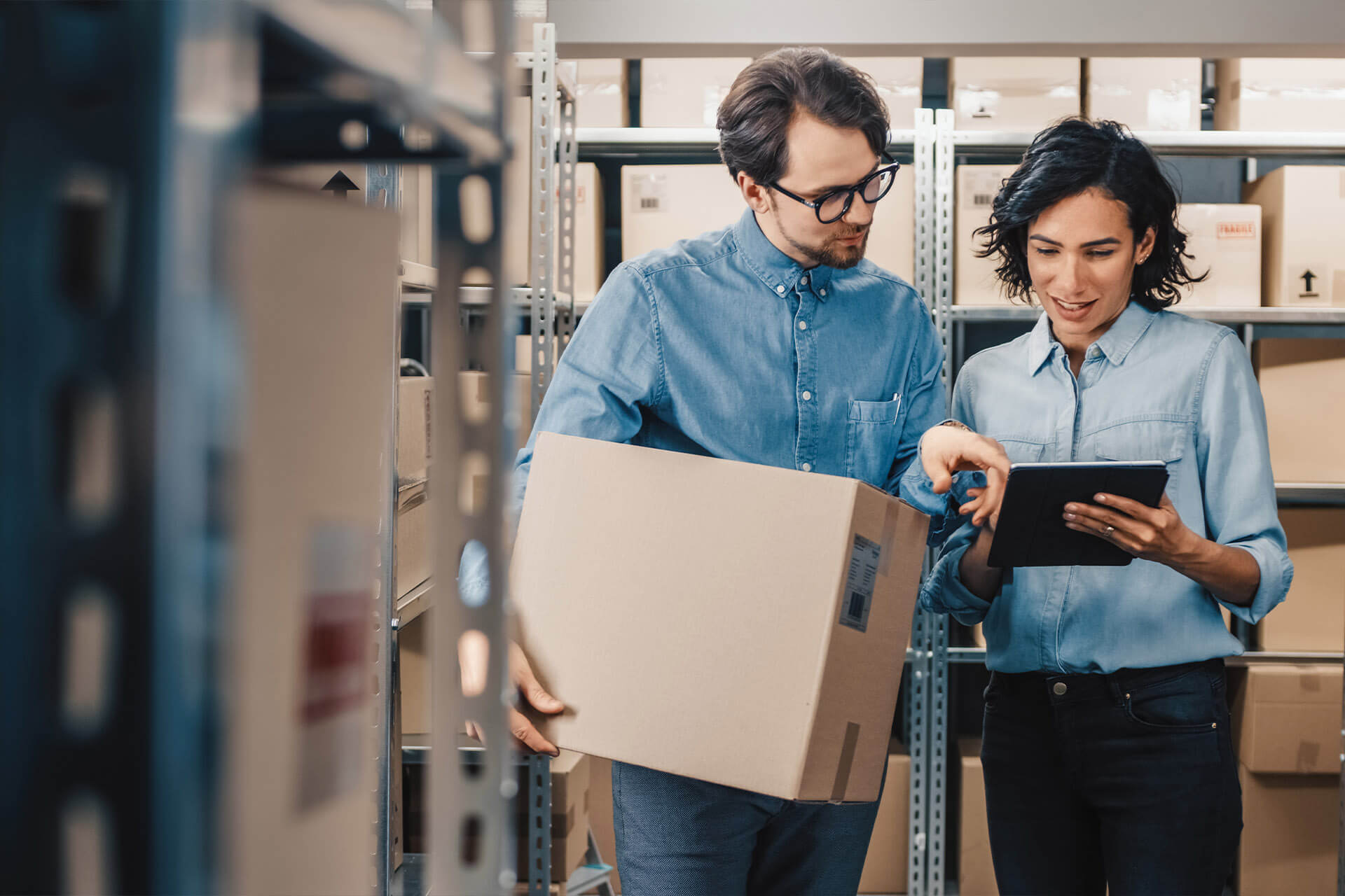 Man and woman standing in room with cardboard boxes on racking, looking at something on a tablet