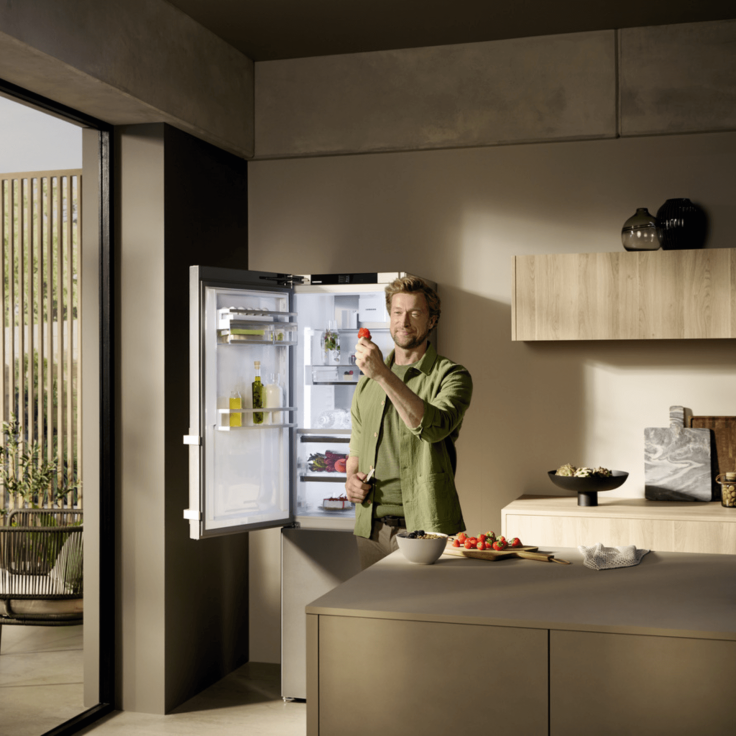 Man standing in front of open fridge freezer admiring a cut strawberry in a bright kitchen.