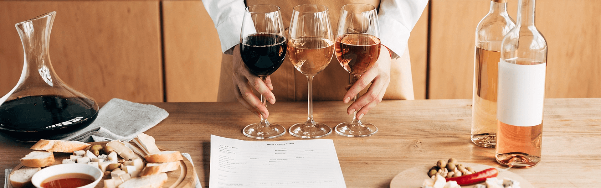 Person in a beige apron standing behind a table with three glasses of wine, red, rosé, and white, arranged in front of them. The table also features a wine decanter, bottles of rosé wine, tasting notes, and platters with bread, cheese, nuts, and honey for pairing.