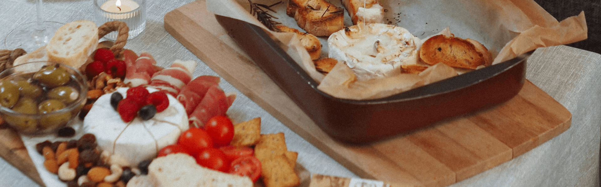 Close-up of a table set with platters of bread and melted cheese, cream cheese poppers with herbs, and a mixed charcuterie board with meats, olives, cheese, and berries, alongside glasses of wine