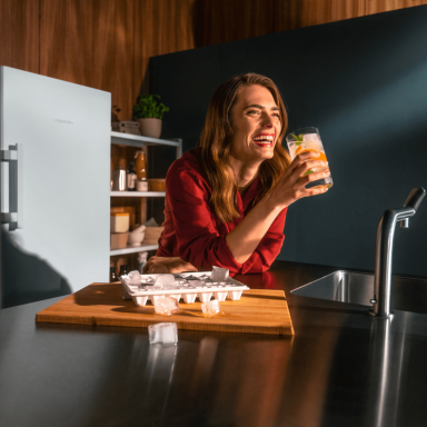 Smiling woman holding a cold beverage whilst she leans on a counter with a Liebherr appliance in the background and an icecube tray next to her with ice popping out of the tray