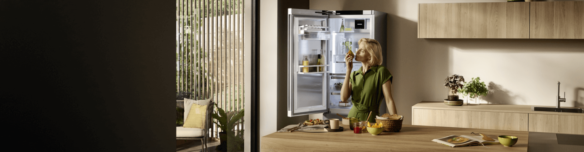 Woman smelling fruit with the open door fridge freezer behind her whilst she prepares a fruit bowl