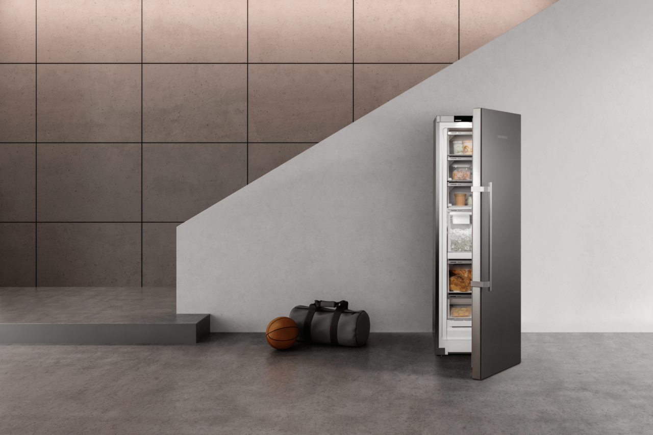 Open door freezer showing frozen food and an ice maker with a gym bag and a basketball next to the freezer. In a modern hallway.