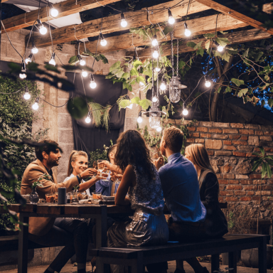 Summer hosting Friends gathered around an outdoor dining table beneath warm lights, enjoying drinks and conversation on a summer evening.
