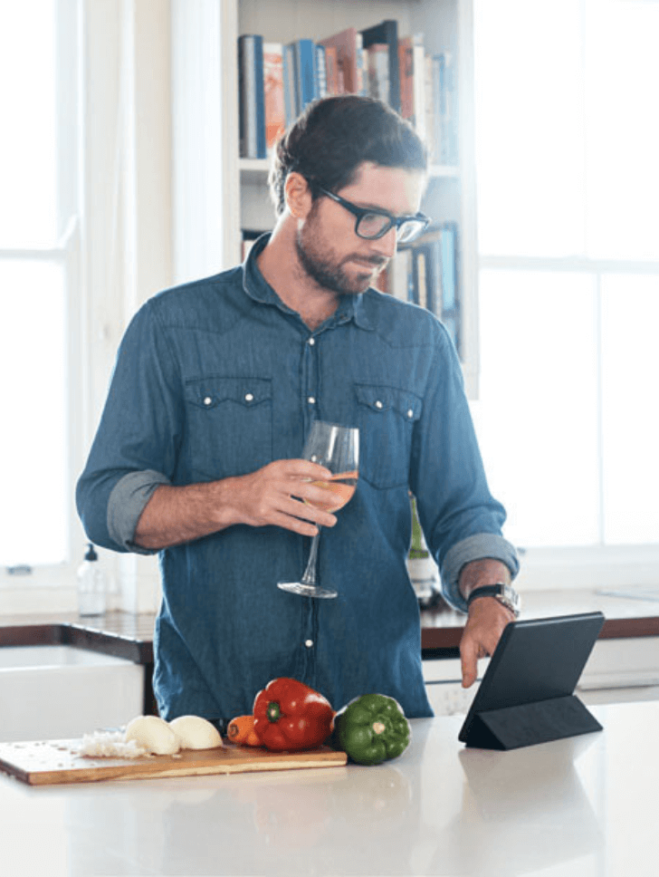 Man in the kitchen holding wine whilst tapping on his tablet