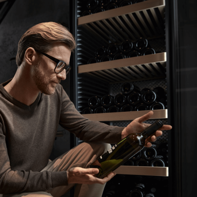 Man looking at a wine bottle with an open door wine fridge in the background