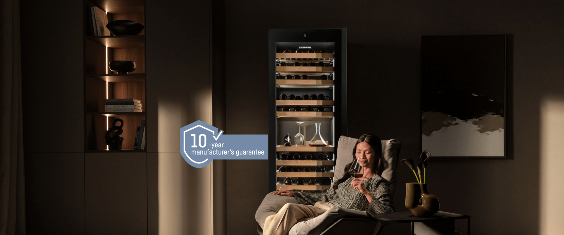 WPgbi 5283 wine fridge with a woman enjoying a glass of wine Woman sitting in a chair holding a glass of red wine in front of a three-zone Liebherr wine fridge, with a blue 10-year guarantee badge displayed on the image.