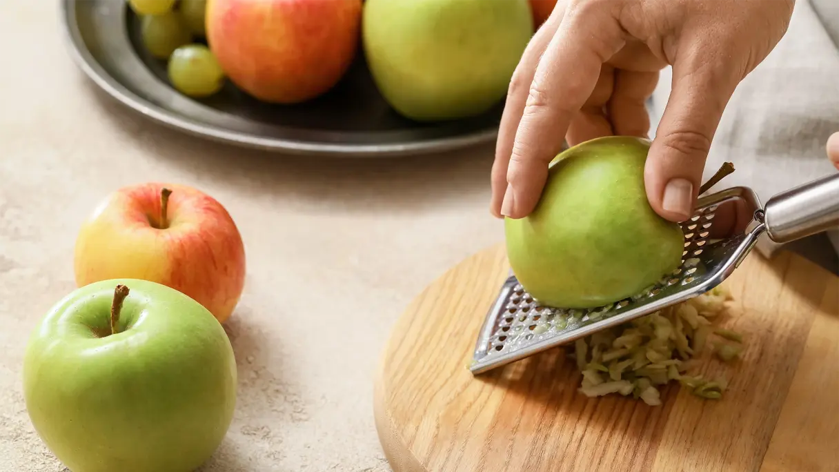 Eine Hand reibt einen Apfel über eine Reibe, daneben liegen weitere Äpfel auf einer Küchenarbeitsplatte.
