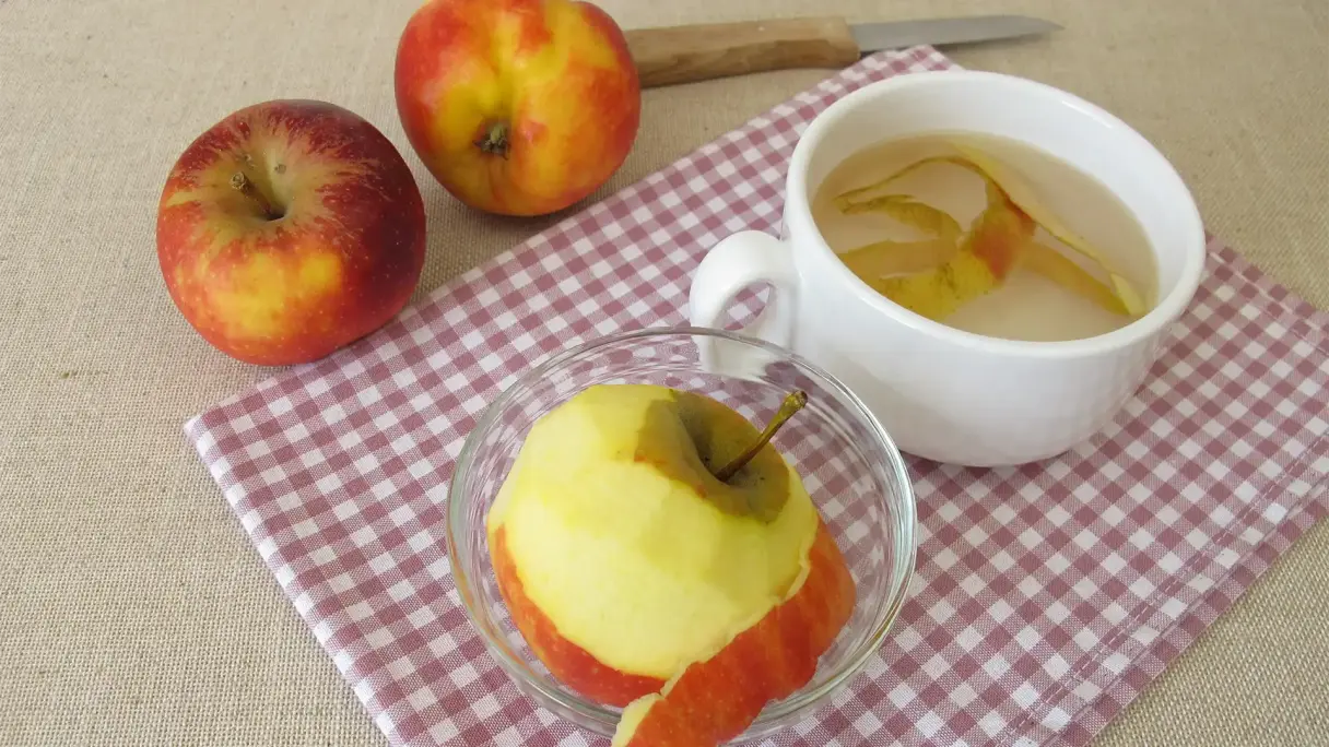 Ein fast geschälter Apfel auf einem rot-weiß karierten Küchentuch, neben einer Tasse mit Wasser und der Apfelschale. Daneben zwei weitere Äpfel.