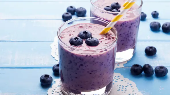 Zwei Gläser mit lila Blaubeer-Smoothie stehen auf einem himmelblauen Tisch. Auf dem Smoothie selbst und auch auf dem Tisch wurden ein paar Blaubeeren verstreut. In den Smoothie-Gläsern steckt jeweils ein weiß-gelb-gestreifter Trinkhalm.
