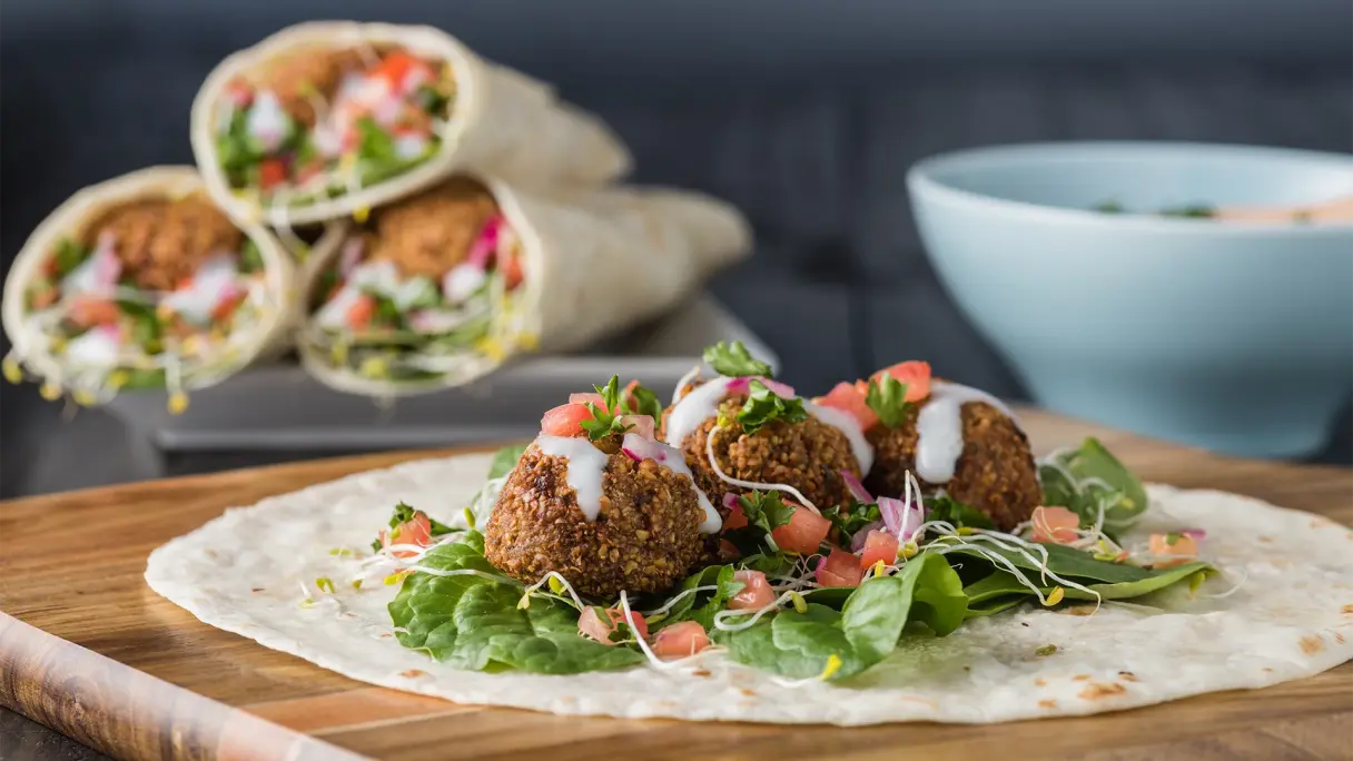 Fast Good: Frischer Falafel-Wrap Fladenbrot mit knusprigen Falafelbällchen, frischem Blattgemüse, Tomatenwürfeln und Sauce auf einem Holzbrett; dahinter angerichtete Wraps und eine Schüssel.