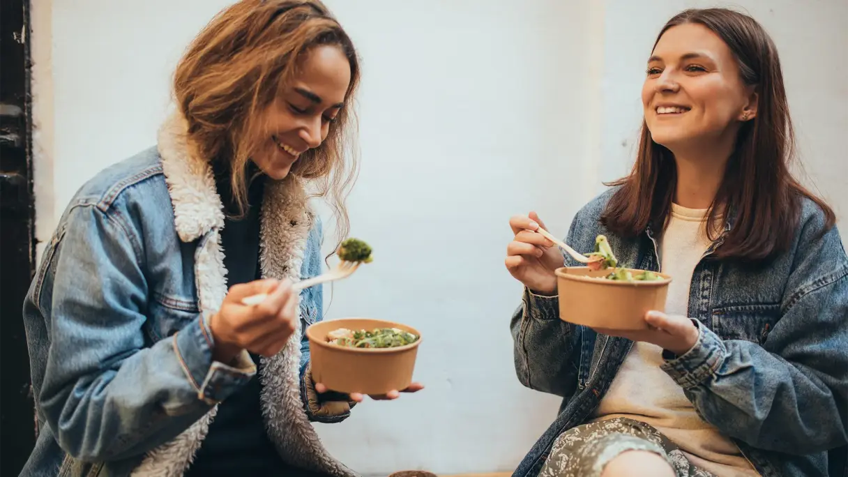 Fast Good: Gemeinsame Pause mit gesunden Bowls Zwei Personen sitzen entspannt zusammen und essen frische Bowls aus braunen Pappschalen; Freizeitmoment in gemütlicher Umgebung.