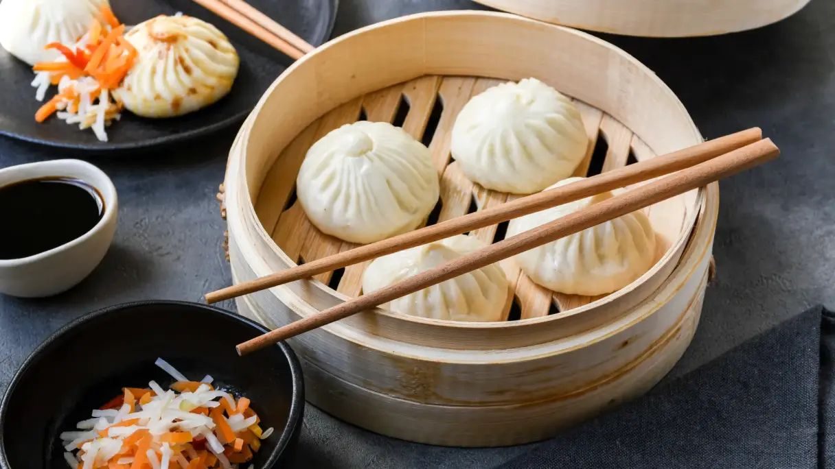 Steamed Asian dumplings such as momos and bao buns arranged in a bamboo steamer, with dipping sauces and chopsticks beside.