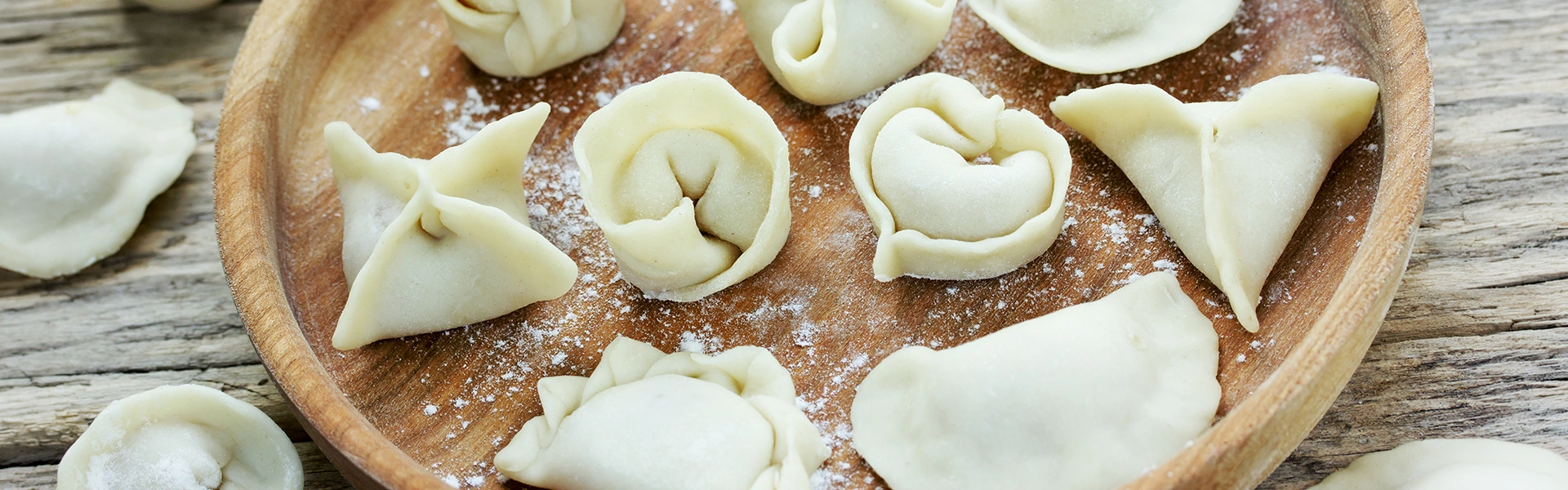 Uncooked filled dumplings in various folding styles arranged on a floured wooden plate, representing international dumpling traditions.