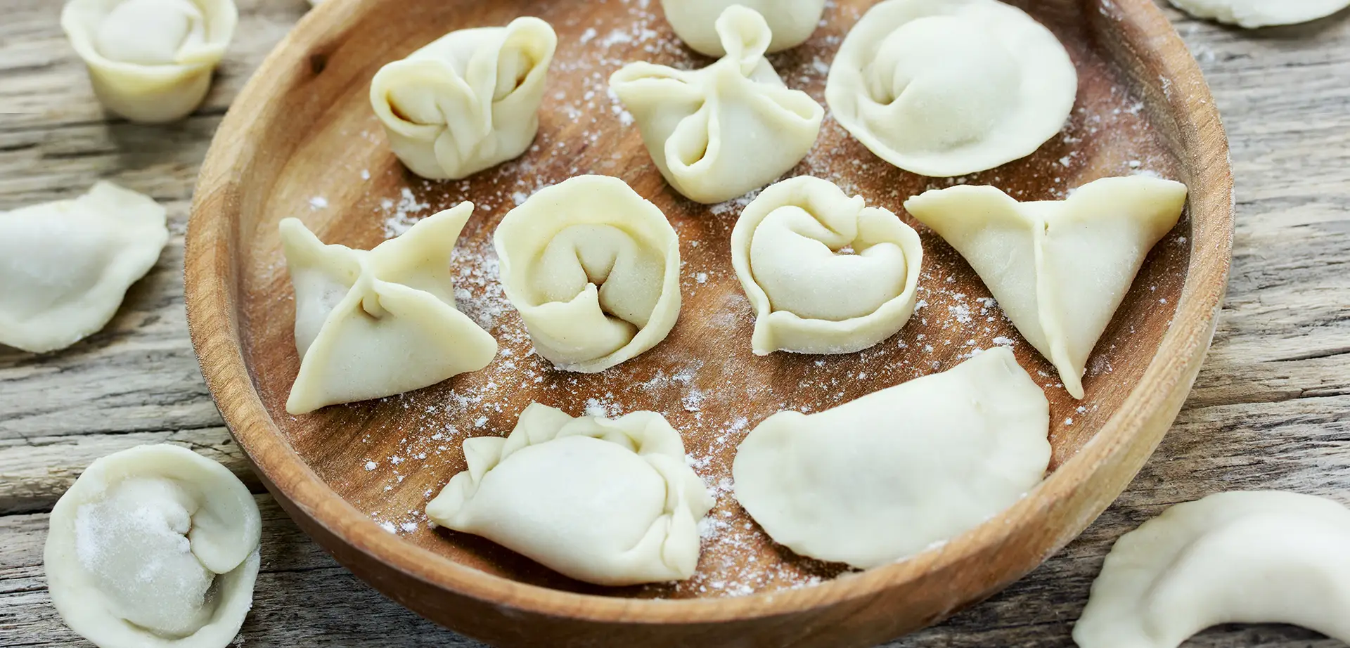 Uncooked filled dumplings in various folding styles arranged on a floured wooden plate, representing international dumpling traditions.