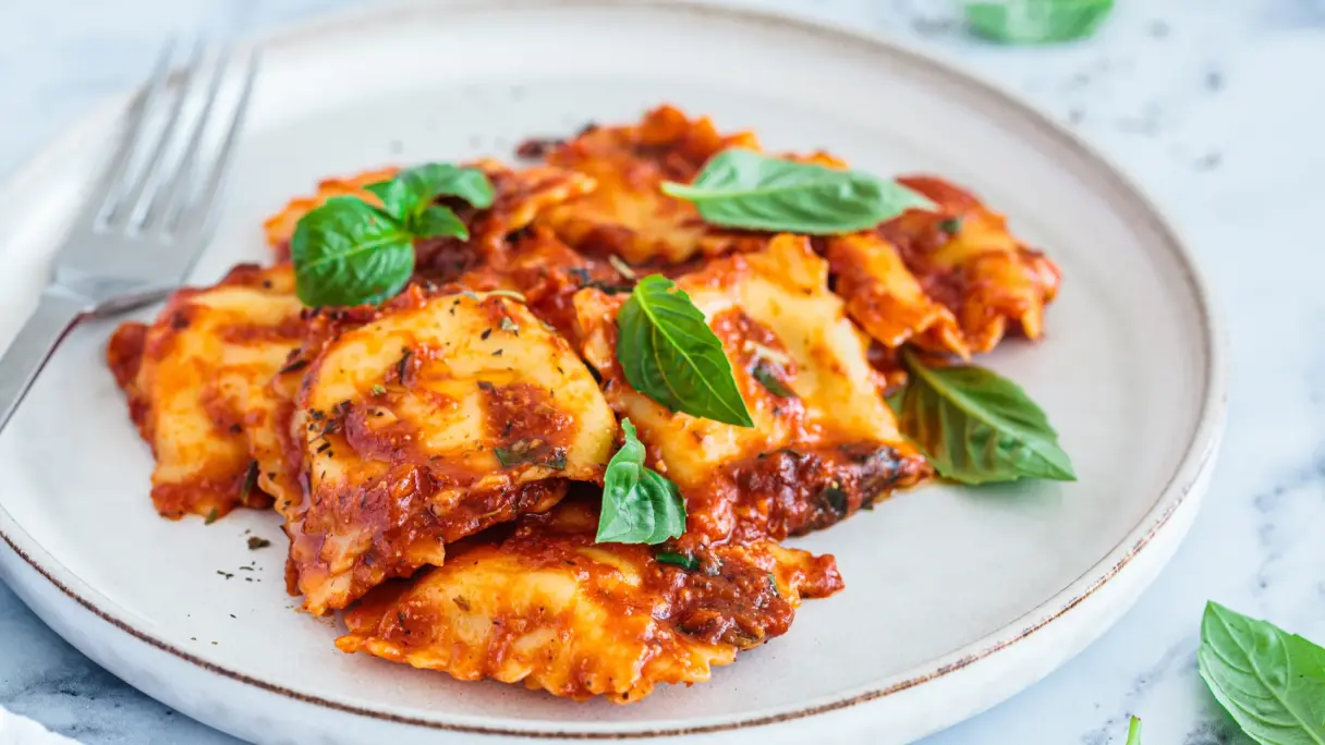 Italian ravioli served on a light plate with rich tomato sauce, topped with fresh basil leaves.