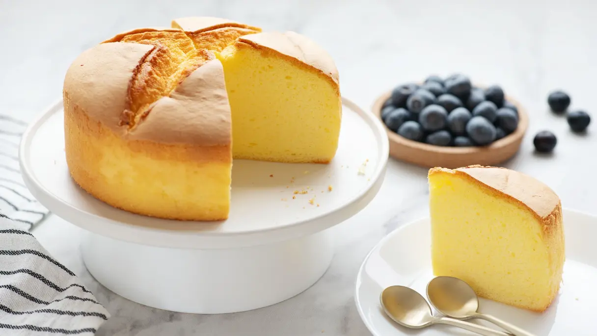 Japanese cheesecake with a slice cut out. A fluffy Japanese cheesecake on a white cake stand, with a slice cut out, and blueberries beside it.