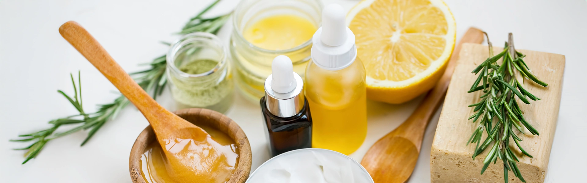 Natürliche Kosmetikzutaten auf weißem Hintergrund: Honig in einem Holzschälchen mit Holzlöffel, Öl in kleinen Glasfläschchen, Heilerde, Creme, Salz, Rosmarinzweige, Seife und Zitrone.