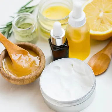 Natürliche Kosmetikzutaten auf weißem Hintergrund: Honig in einem Holzschälchen mit Holzlöffel, Öl in kleinen Glasfläschchen, Heilerde, Creme, Salz, Rosmarinzweige, Seife und Zitrone.