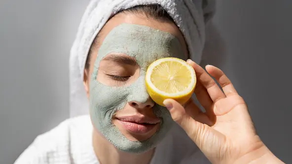 Eine junge Frau lächelt mit geschlossenen Augen und genießt ihre natürliche Gesichtsmaske. Sie trägt ein weißes Handtuch auf dem Kopf und einen weißen Bademantel und hält eine halbe Zitrone vor ihrem linken Auge.