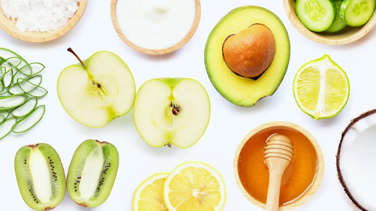 Natürliche Inhaltsstoffe für selbstgemachte Hautpflege: Holzschälchen mit Salz, Aloe Vera, halbierte Kiwi, Joghurt-Schale, halbierter Apfel, Zitronenscheiben, Avocado, Gurkenscheiben im Holzschälchen, Limette, Honig und Kokosnuss auf weißem Hintergrund.