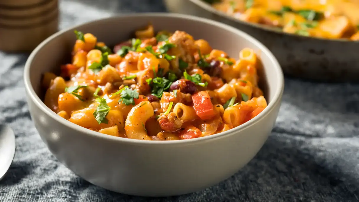 Mac ’n’ Cheese mit Gemüse kombiniert Schüssel mit cremigem Mac ’n’ Cheese, kombiniert mit Gemüsewürfeln und frischen Kräutern; im Hintergrund eine Pfanne mit weiterer Portion.