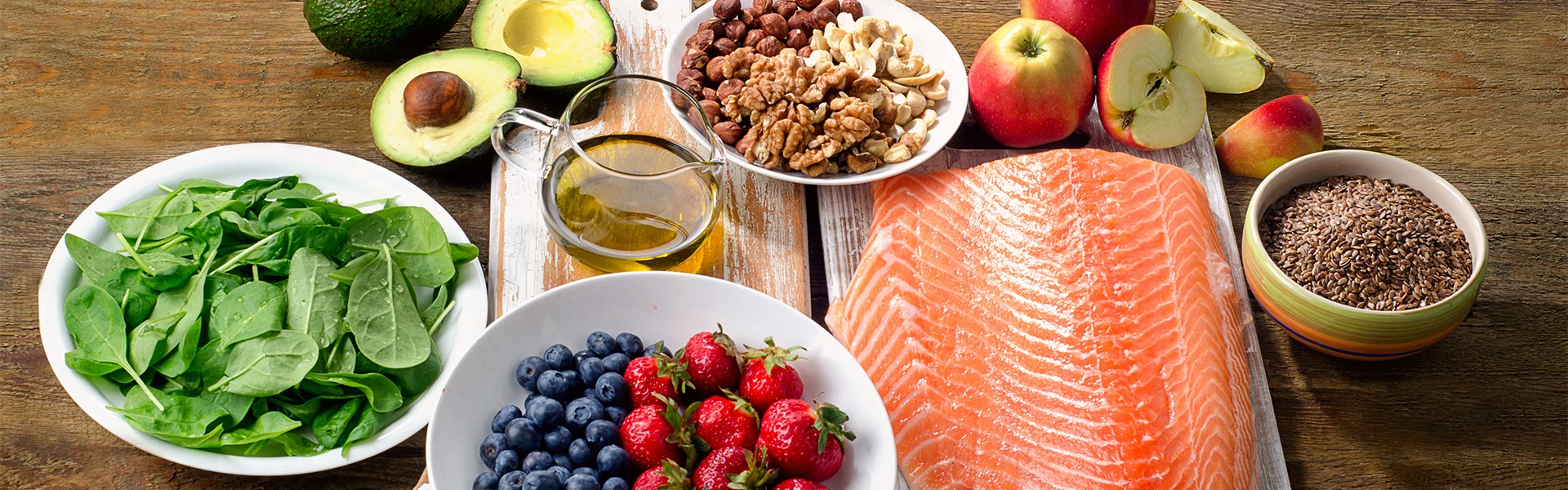 Verschiedene Lebensmittel wie Lachs, Avocado, Beeren, Nüsse, Spinat, Leinsamen und Äpfel liegen auf Holzküchenbrettern auf einem dunklen Holzuntergrund.