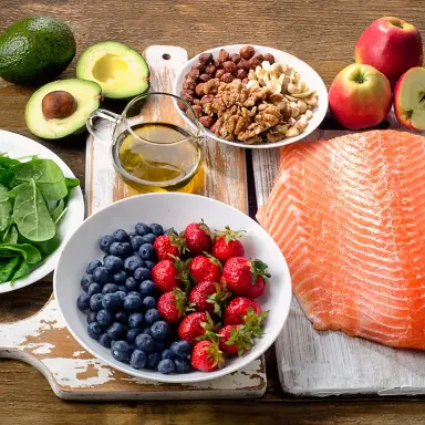 Verschiedene Lebensmittel wie Lachs, Avocado, Beeren, Nüsse, Spinat, Leinsamen und Äpfel liegen auf Holzküchenbrettern auf einem dunklen Holzuntergrund.