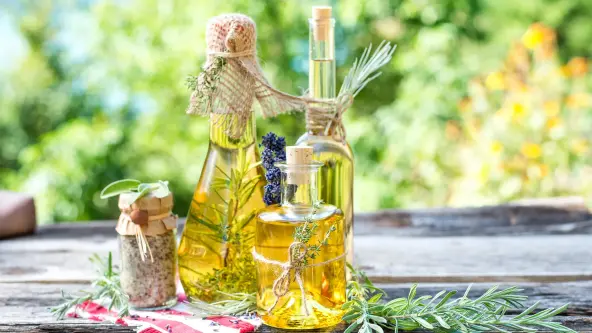 Drei selbst angesetzte Kräuteröle in unterschiedlichen Flaschen stehen neben einem kleinen Glas Kräutersalz auf einem rot-weiß karierten Deckchen auf einem Holztisch im Freien. Drumherum liegen Rosmarinzweige.