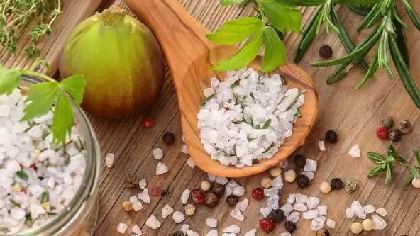 Auf einem Holztisch steht ein Glas mit grobem Salz und Kräutern. Daneben liegt ein Löffel mit diesem grobem Kräutersalz. Drumherum sind frische Kräuter wie Rosmarin und Thymian zu sehen.