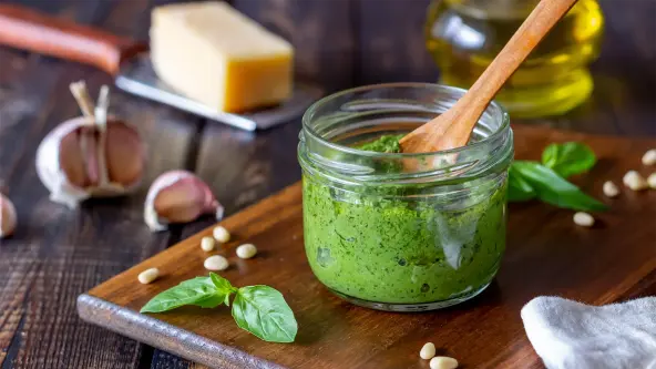 Ein kleines Glas mit frischer grüner Pesto Genovese, in dem ein Holzlöffel steckt, steht auf einem Holzbrett auf einem dunklen Holztisch. Drumherum liegen die Zutaten wie Parmesan, Olivenöl, Knoblauch und Pinienkerne.