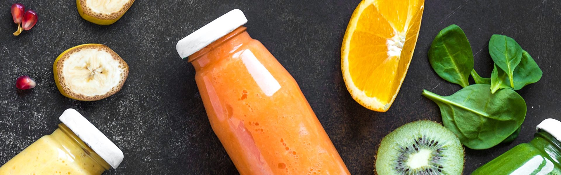 Ein orangener, ein grüner und ein gelber Smoothie liegen in Glasflaschen auf einem dunklen Untergrund. Spinatblätter, ein Stück Orange, eine halbe Kiwi, Bananenscheiben und Granatapfelkerne liegen darum herum verteilt.