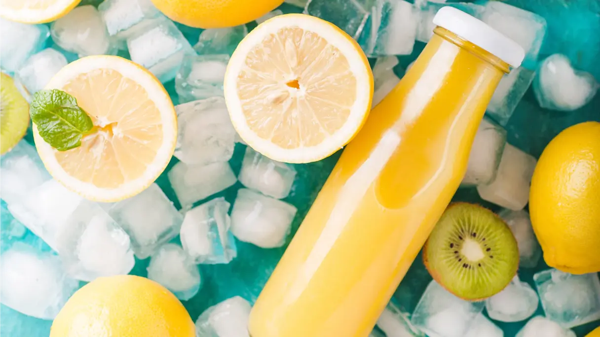 Eine Glasflasche voll gelbem Smoothie liegt auf Eiswürfeln auf einem türkisen Untergrund. Zitronen- und Kiwihälften umrahmen die Flasche.