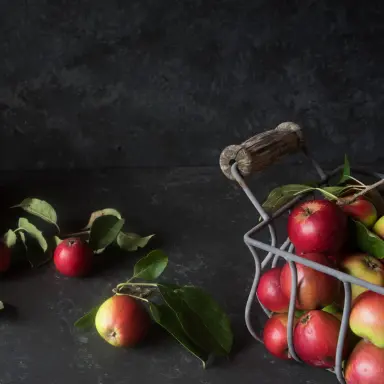Rot-gelbe Äpfel mit Stiel und Blättern liegen teils in einem Metallkorb, teils auf einem anthrazitfarbenen Untergrund.