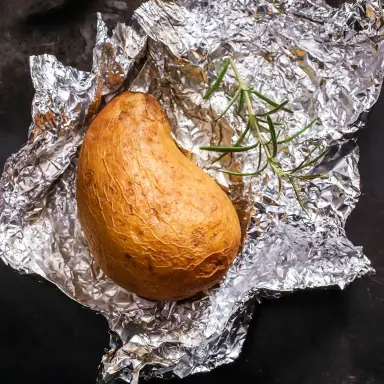 Kartoffel in Alufolie, daneben frischer Rosmarin und eine Gabel auf schwarzem Hintergrund.