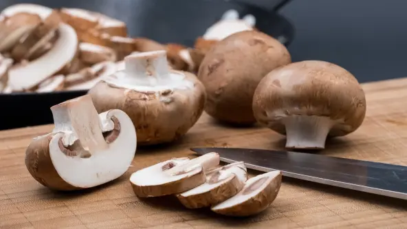 Auf einem Schneidebrett liegen ein Messer und ein in Scheiben geschnittener Champignon sowie drei braune Champignons am Stück.