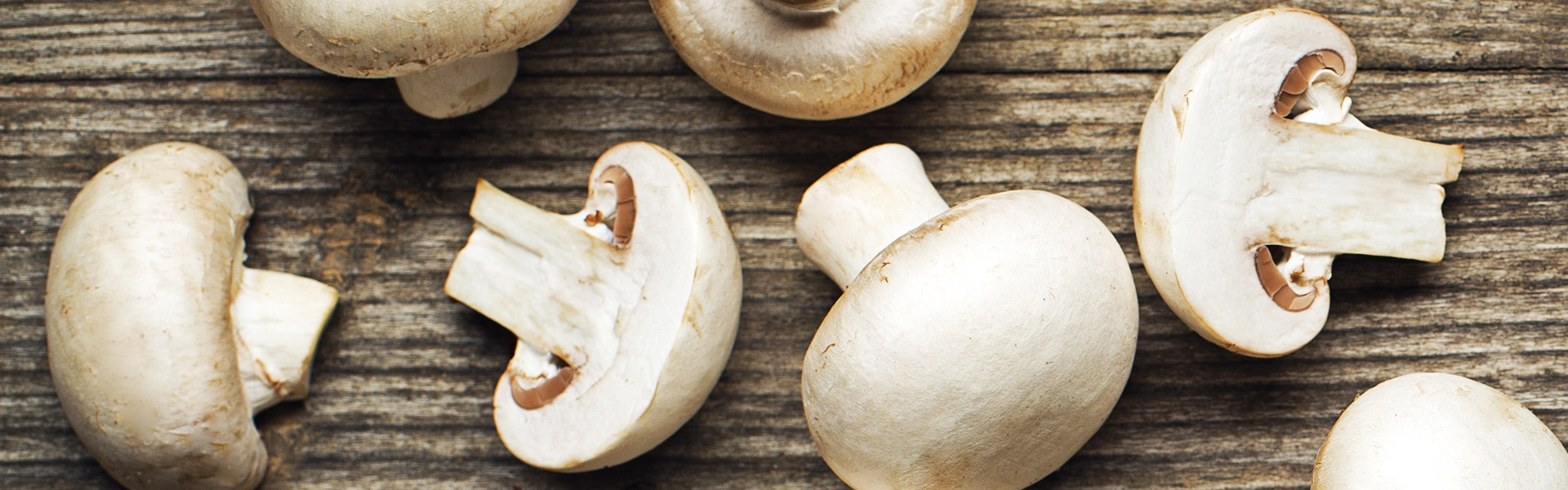 Obenansicht auf ganze und halbe weiße Champignons, die auf einem braunen Holzuntergrund liegen.