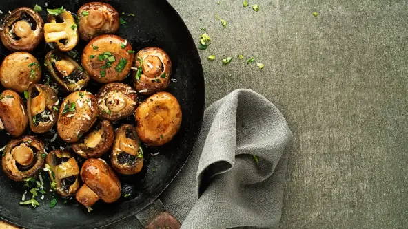 Obenansicht auf eine Pilzpfanne mit Kräutern. Die Champignons sind knusprig braun und wurden mit Kräutern bestreut. Die Pfanne steht auf einem grauen Untergrund.