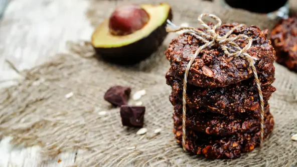 Vier dunkelbraune Schokoladen-Kekse sind übereinandergestapelt und mit einem Faden verschnürt. Daneben liegt eine halbe Avocado sowie ein paar Schokoladen-Stückchen.