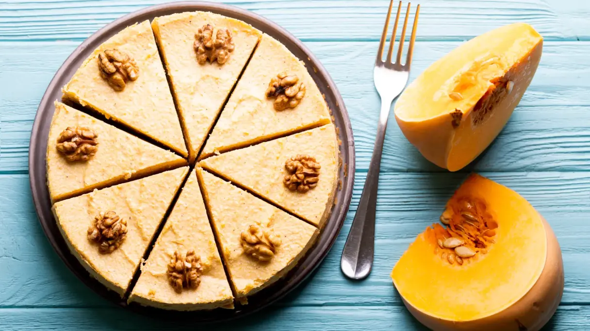 Obenansicht eines Pumpkin Pies – eines Kuchens mit Kürbis als Zutat. Der Kuchen ist in 8 Stücke geschnitten und hat eine hellbraune Farbe. Neben dem Kuchen liegen zwei Viertel von einem Butternut-Kürbis.