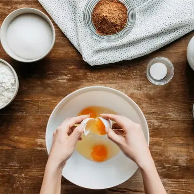 Ein weißes Ei wird über einer Schüssel aufgeschlagen. Eigelb und Eiweiß sind schon zu sehen. In der Schüssel befinden sich bereits zwei aufgeschlagene Eier.
