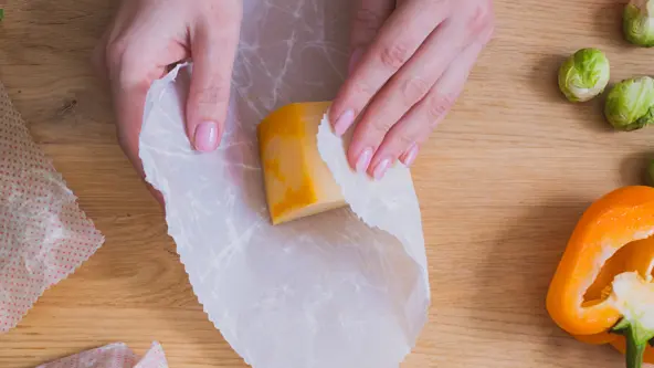 Einfrieren mit Wachstüchern Hände, die einen Käse auf einer Holzarbeitsplatte in ein Wachstuch wickeln, daneben Gemüse und weitere Wachstücher.