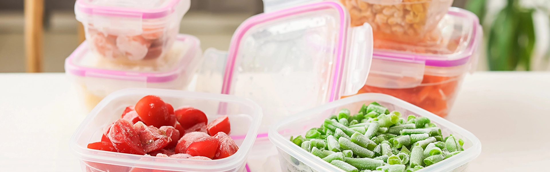 Eine Küchenarbeitsplatte mit verschiedenen Aufbewahrungsboxen, gefüllt mit Gemüse. Im Vordergrund sind zwei geöffnete Behälter mit gefrorenen Tomaten und gefrorenen Bohnen zu sehen.