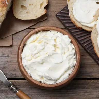Obenansicht auf eine Schüssel Frischkäse auf einem Holztisch. Daneben liegen mit Frischkäse bestrichene Scheiben Weißbrot sowie ein Brotmesser.
