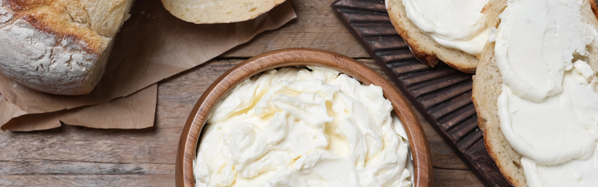Obenansicht auf eine Schüssel Frischkäse auf einem Holztisch. Daneben liegen mit Frischkäse bestrichene Scheiben Weißbrot sowie ein Brotmesser.