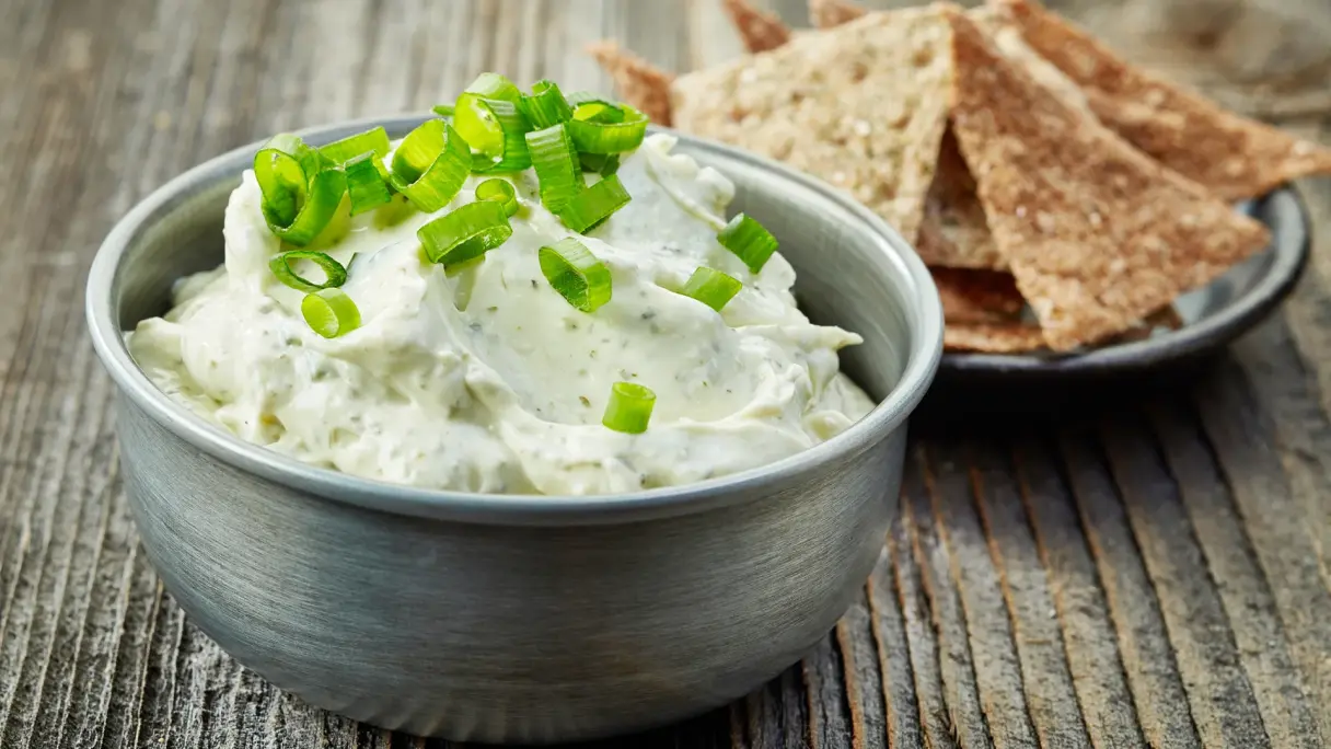 Eine Schüssel Frischkäse mit grünen Zwiebeln und Kräutern steht auf einem Holztisch. Dahinter steht ein Teller mit Knäckebrot.