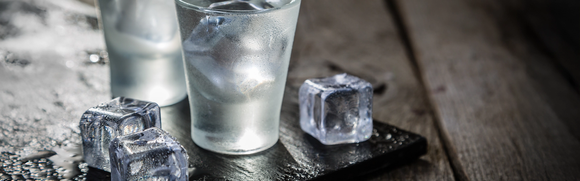 Zwei klare Gläser mit kalter, klarer Flüssigkeit und Eiswürfeln stehen auf einer dunklen Steinplatte auf einem dunklen Holztisch. Daneben liegen weitere Eiswürfel.