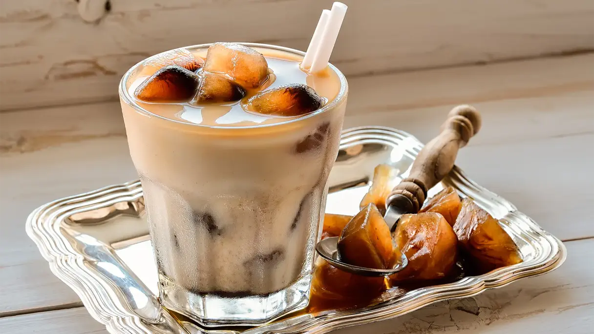 Ein Glas mit Eiskaffee steht auf einem kleinen silbernen Tablett, daneben Kaffeeeiswürfel und ein Löffel auf Holzhintergrund.