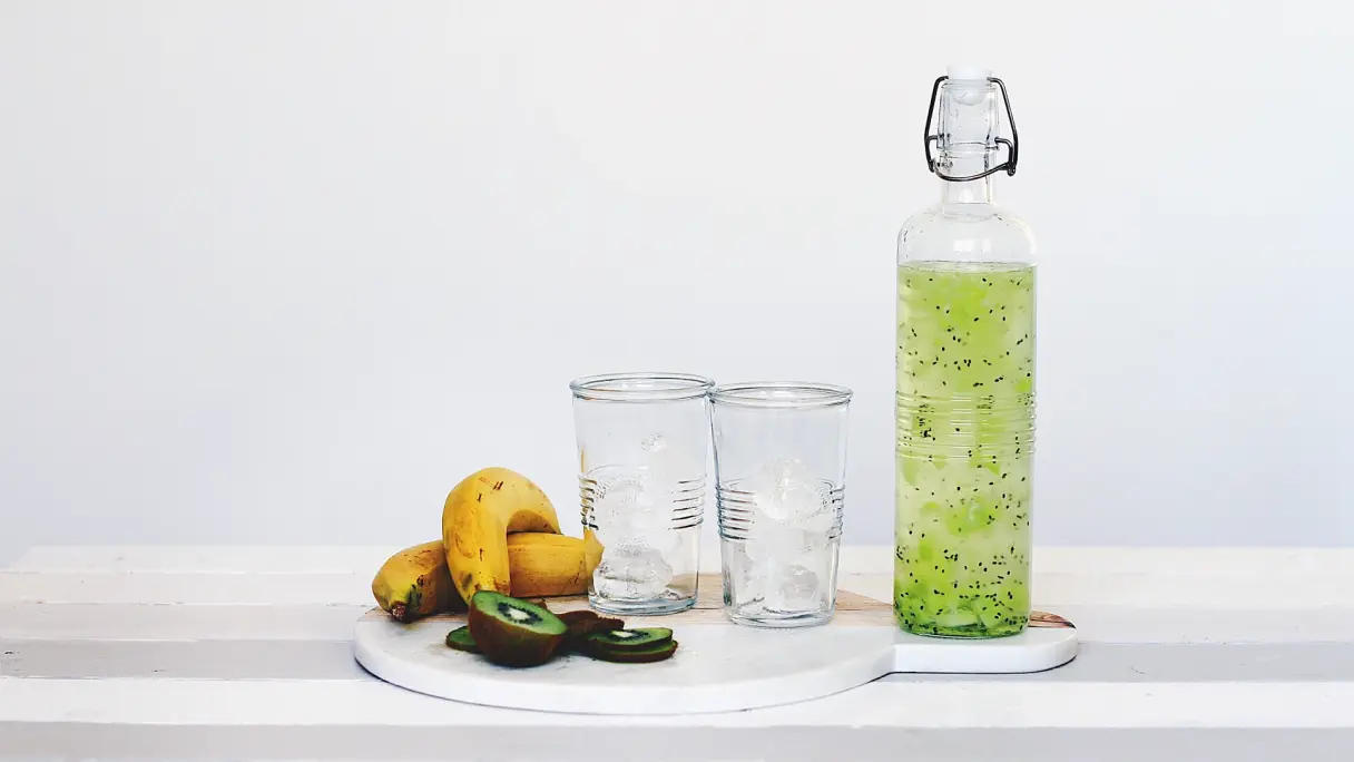 Eine Flasche Detox-Wasser und zwei Gläser stehen auf einem Tisch. Das Wasser ist hellgrün und hat winzige schwarze Kerne darin. Neben den Gläsern liegen Kiwischeiben und zwei Bananen.